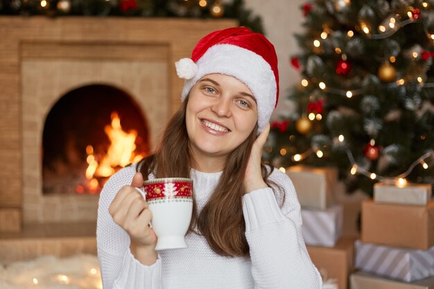Kobieta z kubkiem w dłoniach siedzi w salonie przy kominku, z zębatym uśmiechem, ciesząc się gorącym napojem w przytulnej atmosferze, dziewczyna pozuje w pobliżu choinki i prezentów.