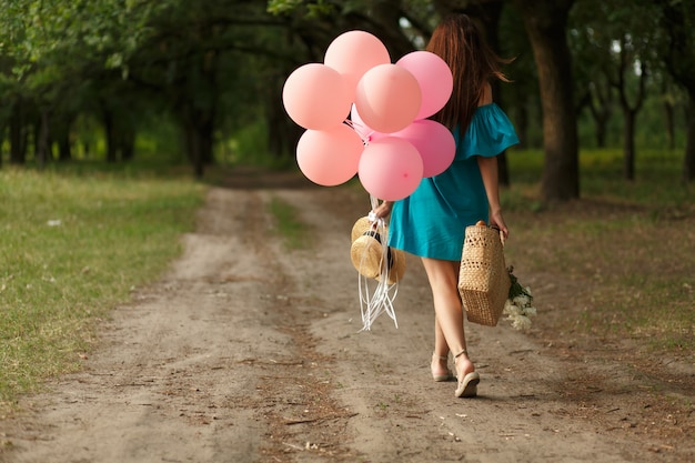 Kobieta Z łozinowym Koszem, Kapeluszem, Różowymi Balonami I Kwiatami Chodzi Na Wiejskiej Drodze