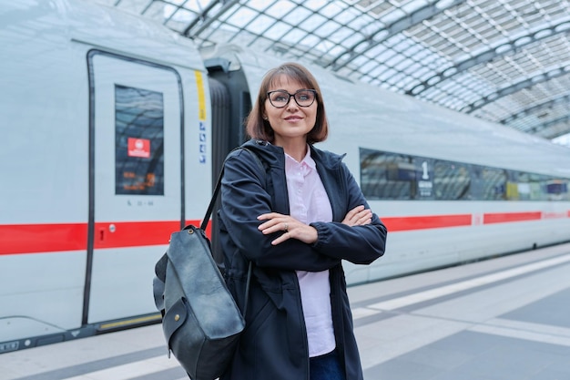 Kobieta z plecakiem czekająca na peronie kolejowym wewnątrz stacji uśmiechnięta kobieta ze skrzyżowanymi ramionami patrząca na kamerę Transport kolejowy Transport pasażerów Podróż podróż podróż ludzi