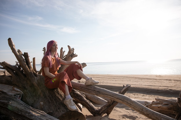 kobieta z różowymi włosami siedzi na drewnianych balach na plaży i cieszy się zachodzącym słońcem