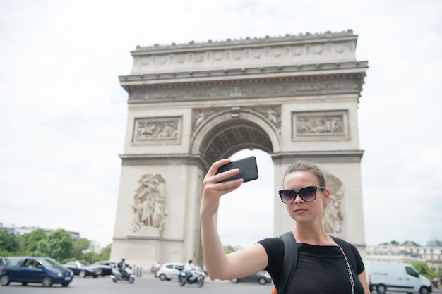 Kobieta z smartphone o łuk pomnik w paryżu, francja. Kobieta zrobić selfie z telefonem w łuk triumfalny. Wakacje i zwiedzanie stolicy Francji. Dziewczyna z modnym wyglądem i zmysłowym pięknem.