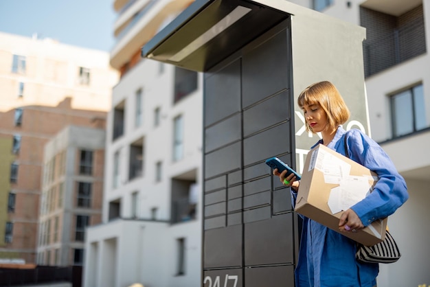 Zdjęcie kobieta z telefonem i paczką w pobliżu automatycznej maszyny pocztowej w dzielnicy mieszkalnej