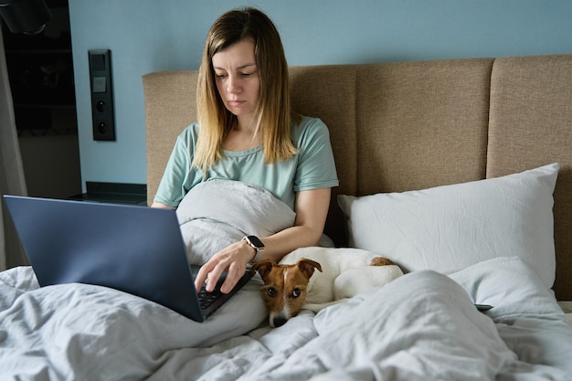 Kobieta z uroczym psem leżącym w łóżku i korzystającym z laptopa rano