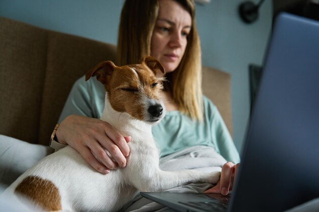 Kobieta z uroczym psem relaksująca się rano w łóżku i korzystająca z laptopa, wygodna praca w domu