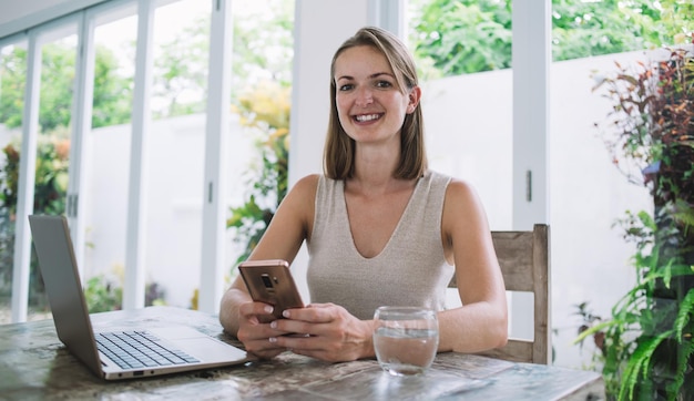 Kobieta z uśmiechem używająca smartfona i patrząca w kamerę przy stole z laptopem i szklanką wody