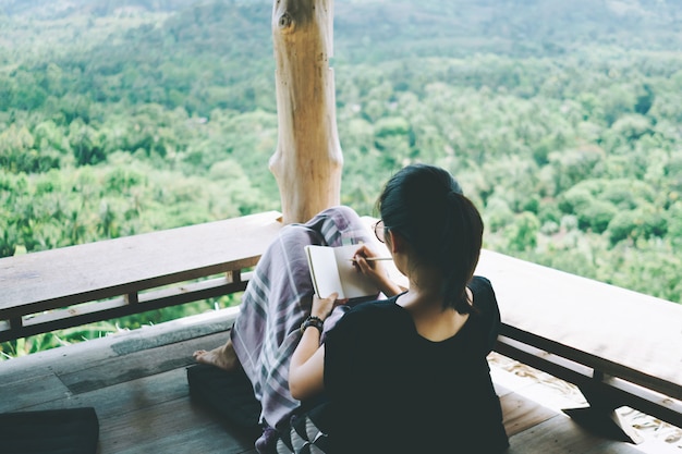 Kobieta zapisuje w małym białym notatniku, aby zrobić notatkę, aby nie zapomnieć, zaplanować lub napisać książkę z pięknym tarasem z widokiem na dom i góry.