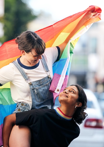 Kobiety lgbtq para i piggyback z flagą dla dumy, miłości i wsparcia na ulicy miasta Różnorodność przyjaciele lesbijki i geje wspólnie świętują tęczową tożsamość wolność i szczęście praw człowieka