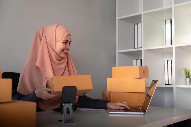 Kobiety muzułmańskie sprzedające online w domu z pudełkiem Sprzedające online z pudełkiem do przyjmowania zamówienia od klienta pomysł biznesowy MŚP Dostawa paczek muzułmańska kobieta pracująca ze smartfonem i laptopem w domu.