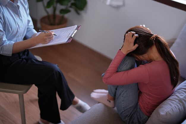Kobiety z Azji cierpiące na stres i bóle głowy z powodu niepokoju, podczas gdy psycholog bada problemy ze zdrowiem psychicznym i pisze notatki na papierze do doradztwa w zakresie terapii zdrowia psychicznego