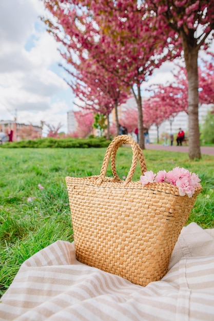 Koc piknikowy ze słomkowym kapeluszem i torbą na zielonej trawie pokrytej różowymi kwiatami sakura kopia przestrzeń