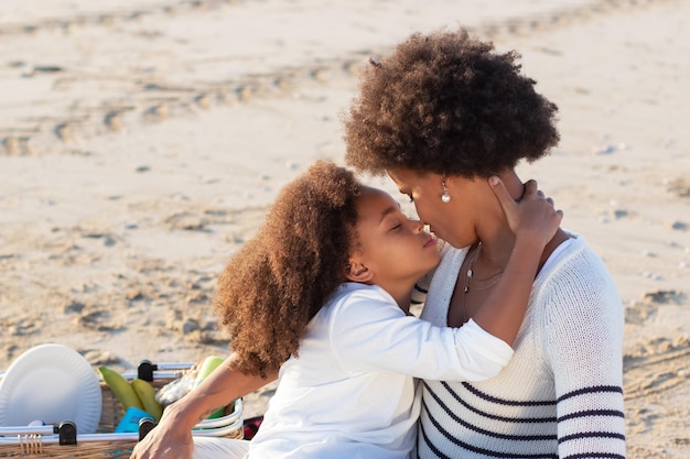 Kochająca Afroamerykańska Rodzina Na Pikniku Na Plaży. Matka I Córka W Ubranie Siedzi Na Kocu, Przytulanie. Rodzina, Relaks, Koncepcja Natury