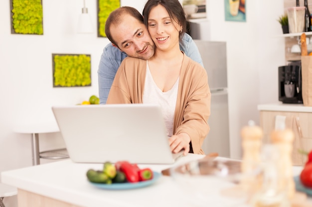 Kochająca Para Pracuje Na Laptopie W Kuchni Ze świeżymi Warzywami Na Stole. Szczęśliwa Kochająca Wesoła Romantyczna Zakochana Para W Domu Przy Użyciu Nowoczesnej Technologii Bezprzewodowego Internetu Wi-fi