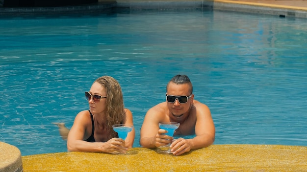 Kochająca się para przytula się i całuje pijąc niebieski koktajl alkoholowy alkohol w basenie w hotelu na zewnątrz Portret kaukaskiego mężczyzny i kobiety Kreatywne fryzury Kulturysta strój kąpielowy Okulary przeciwsłoneczne