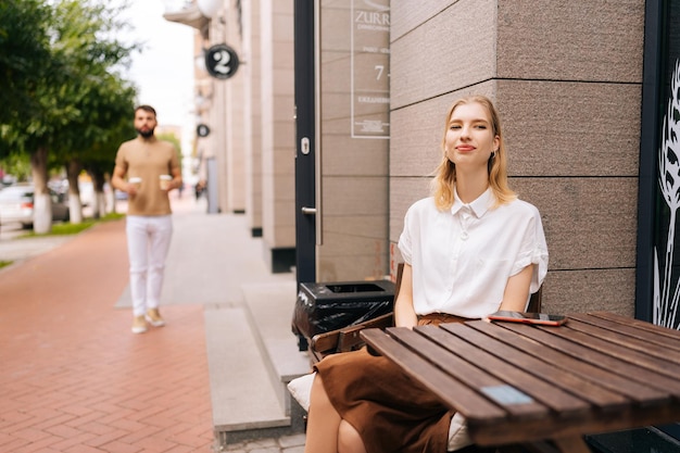 Kochający młody człowiek przynoszący kawę uroczej blondynce siedzącej przy stole w ulicznej kawiarni w letni dzień