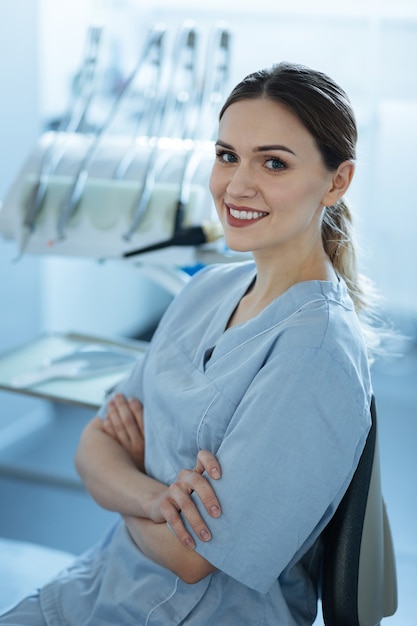 Kocham Moje Miejsce Pracy. Dość Młoda Dentystka Pozuje Przed Aparatem Dentystycznym W Swoim Gabinecie I Składa Ręce Na Piersi, Uśmiechając Się Szeroko