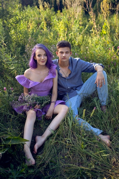 Zdjęcie kochankowie to piękny facet z dziewczyną z fioletowymi włosami i bukietem kwiatów siedzącym na trawie latem