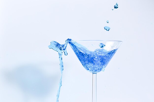 Koktajl z niebieskim płynem w szkle. Szklanka z niebieską wodą zalewaną płynem z rozpryskami i kroplami. Szklanka martini z alkoholem z plamami na białym tle. Koncepcja napoju orzeźwiającego.