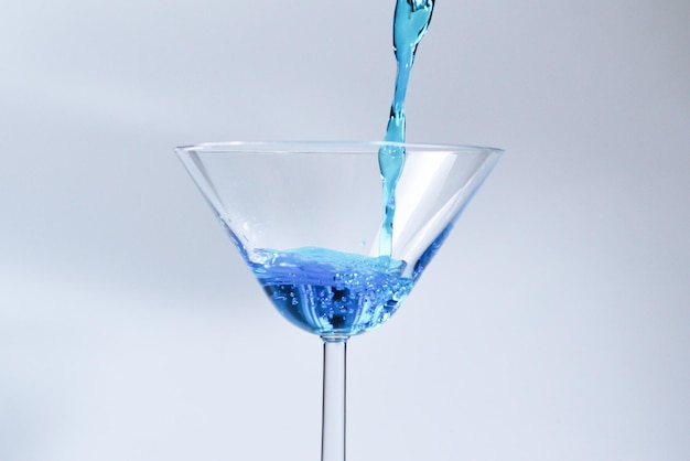 Koktajl z niebieskim płynem w szkle Szkło z niebieską wodą wlewającą się z płynem z plamami i kroplami Napełnianie szkła Martini alkoholem z plamami na białym tle Koncepcja napoju orzeźwiającego