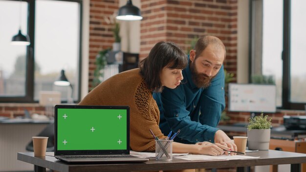 Koledzy pracujący i posiadający laptopa z zielonym ekranem na wyświetlaczu w biurze startowym. Ludzie biznesu projektujący strategię projektu, mając jednocześnie klucz chroma i szablon makiety na białym tle.