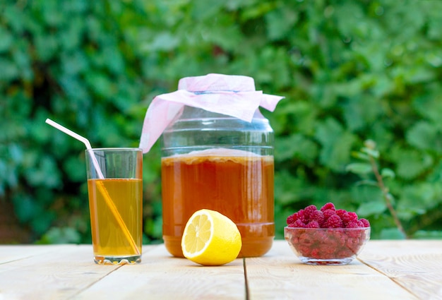 Kombucha w słoiku na lekkim drewnianym stole na tle nieba, szklanka wypełniona Kombuchą z malinami.