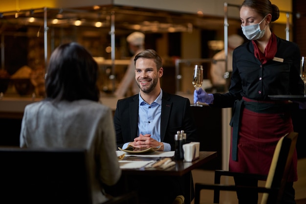 Kompetentna kelnerka trzymająca w lewej ręce kieliszek z napojem i stawiająca go na stole między dwiema osobami