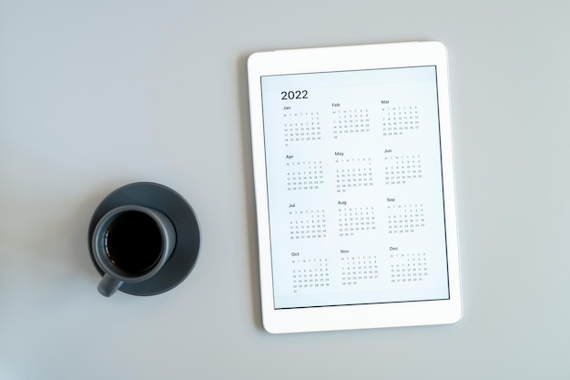 Komputer typu Tablet z otwartą aplikacją kalendarza na rok 2022 i filiżanką herbaty lub kawy na szarym tle. koncepcja biznesu lub lista celów z wykorzystaniem technologii. widok z góry, układ płaski