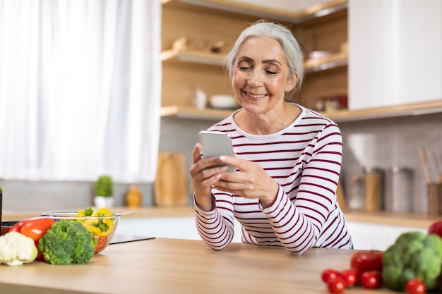 Komunikacja mobilna portret uśmiechniętej starszej pani za pomocą smartfona w kuchni