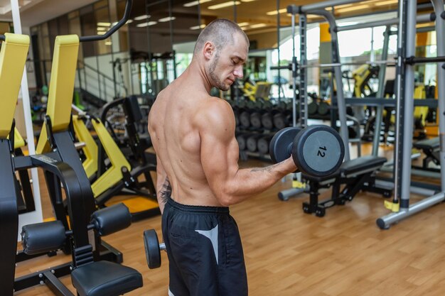 Koncentruje się na treningu muskularny szczupły mężczyzna ćwiczący z hantlami w nowoczesnej siłowni. Pompowanie bicepsów