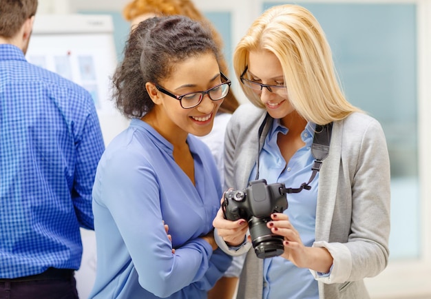 Koncepcja Biznesowa, Fotograficzna I Startupowa - Dwie Uśmiechnięte Młode Kobiety Patrzące Na Aparat Cyfrowy W Biurze