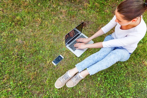 Koncepcja biznesowa freelancer. Młoda kobieta siedzi na zielonym trawniku w parku miejskim pracy na komputerze typu laptop. Styl życia autentyczny szczery studentka studiująca na zewnątrz. Biuro mobilne