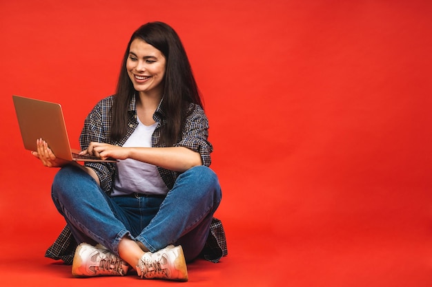 Koncepcja biznesowa Portret szczęśliwej brunetki kobiety w przypadkowym siedzeniu na podłodze w pozie lotosu i trzymającego laptopa odizolowanego na czerwonym tle