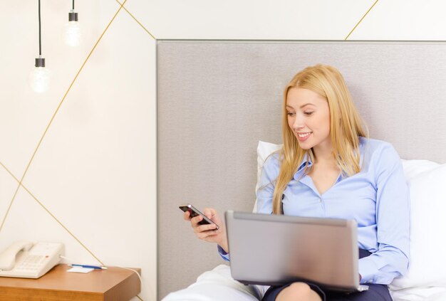 koncepcja biznesowa, technologia, internet i hotel - szczęśliwa bizneswoman ze smartfonem i laptopem leżącym w hotelu w łóżku