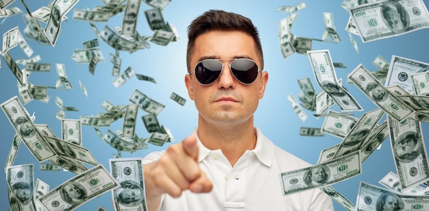 koncepcja biznesu, finansów, gestów i ludzi - twarz latynoskiego mężczyzny w średnim wieku w okularach przeciwsłonecznych, wskazując palcem na ciebie na niebieskim tle ze stertą spadających pieniędzy w dolarach
