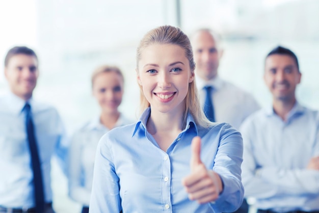 koncepcja biznesu, ludzi, gestów i pracy zespołowej - uśmiechnięta kobieta pokazująca kciuk w górę z grupą biznesmenów w biurze