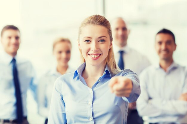 koncepcja biznesu, ludzi i pracy zespołowej - uśmiechnięta kobieta wskazująca palcem na ciebie z grupą biznesmenów w biurze