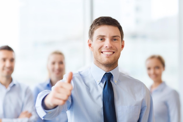 koncepcja biznesu, ludzi i pracy zespołowej - uśmiechnięty biznesmen pokazujący kciuki w górę z grupą biznesmenów w biurze