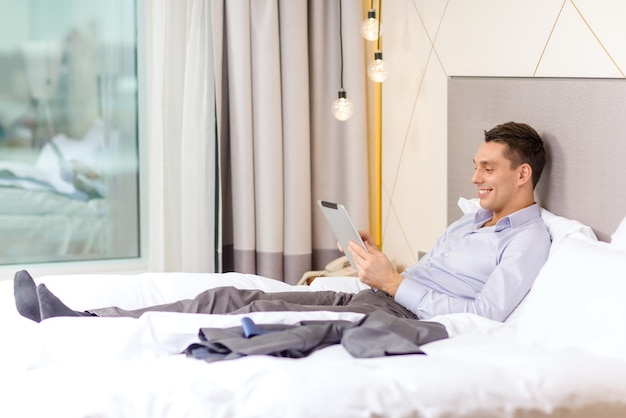 koncepcja biznesu, technologii, internetu i hotelu - szczęśliwy biznesmen z komputerem typu tablet pc leżącym w hotelu w łóżku