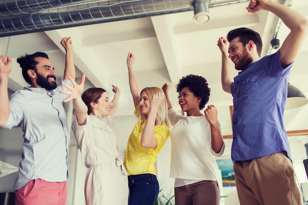 koncepcja biznesu, triumfu, gestu, ludzi i pracy zespołowej - szczęśliwy kreatywny zespół podnoszący ręce do góry i świętujący zwycięstwo w biurze