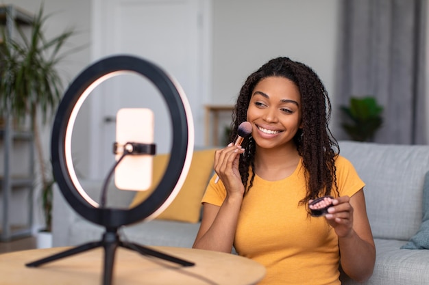 Koncepcja blogowania piękna szczęśliwa czarna dama nagrywa recenzję makijażu za pomocą lampy pierścieniowej i telefonu komórkowego
