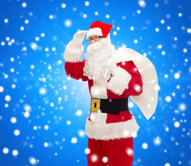 Koncepcja Bożego Narodzenia, Wakacji I Ludzi - Mężczyzna W Stroju świętego Mikołaja Z Torbą Patrzący Daleko Na Niebieskim śnieżnym Tle
