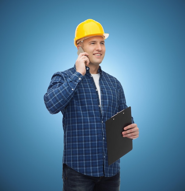 koncepcja budowy, budowy, ludzi, technologii i konserwacji - uśmiechnięty mężczyzna budowniczy lub pracownik fizyczny w kasku ze schowkiem dzwoniącym na smartfona na niebieskim tle