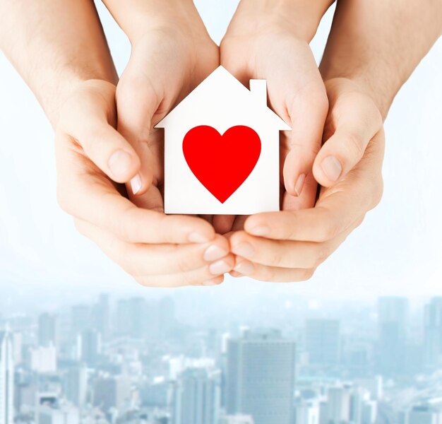 koncepcja charytatywna, nieruchomości i domu rodzinnego - zbliżenie przedstawiające męskie i żeńskie ręce trzymające biały papierowy dom z czerwonym sercem