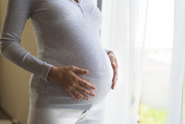 Koncepcja ciąży, kobieta w ciąży stojąca w pobliżu okna z rękami na brzuchu, styl życia w ciąży