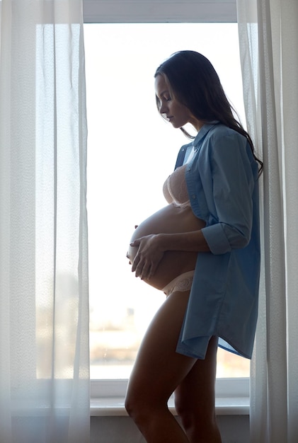 koncepcja ciąży, macierzyństwa, ludzi i oczekiwań - sylwetka szczęśliwej kobiety w ciąży z dużym nagim brzuchem w pobliżu okna w domu