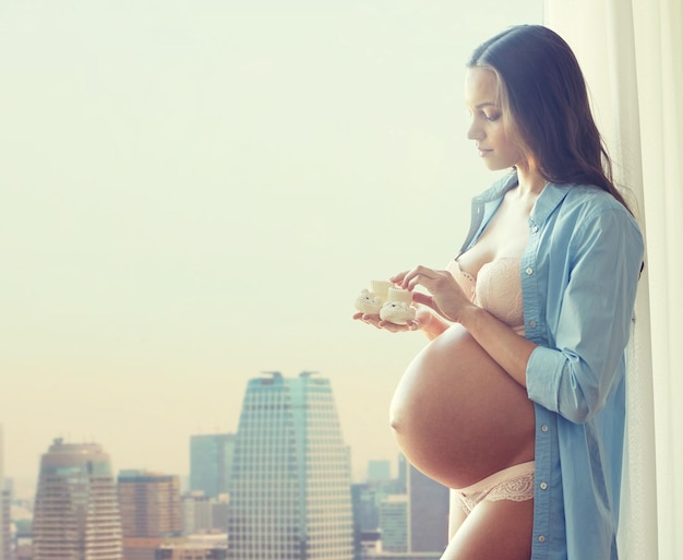 koncepcja ciąży, macierzyństwa, ludzi i oczekiwań - szczęśliwa kobieta w ciąży z dużym nagim brzuchem trzymająca małe dziecięce buciki w domu na tle miasta