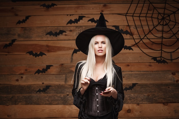 Koncepcja Czarownicy Halloween - Happy Halloween Witch Gospodarstwa Pozowanie Z Smartphone Na Starej Drewnianej ścianie.