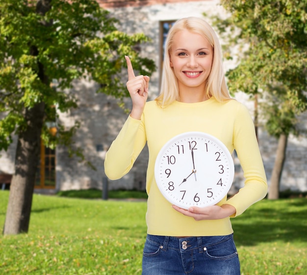 koncepcja czasu, edukacji i ludzi - uśmiechnięta młoda kobieta z zegarem ściennym pokazującym 8 i palcem w górę