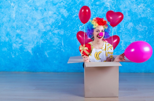 Koncepcja Dnia Głupców 1 kwietnia Zabawna śmiejąca się dziewczyna klaun siedzi w kartonowym pudełku z balonami heliowymi i białą tablicą w ręku prezent urodzinowy