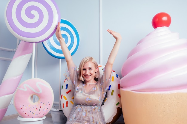 Koncepcja dosładzania. piękna młoda blondynka Barbie słodka kobieta cukiernik model idealny makijaż siedzący obok lizaka i fałszywe słodycze słodycze pączek cukierki ogromne pączki ciasto tło w studio