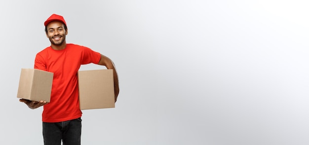 Koncepcja dostawy Portret szczęśliwy African American człowiek dostawy w czerwonym suknem trzyma pakiet pudełko na szarym tle studio Kopia przestrzeń